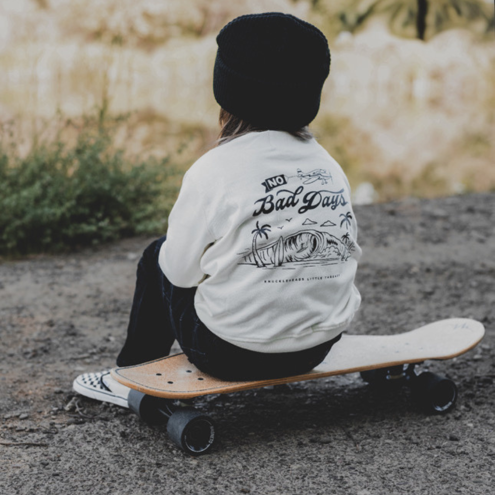 Knuckleheads Off-White Infant Crew Neck for kids, surf-inspired design in lightweight, breathable 100% cotton fabric, displayed against a laid-back, coastal-themed background. Available in sizes from 12 months to 8 years, this top is perfect for casual playdates, beach days, or park outings. Shown with durable, easy-to-wash fabric for stylish, everyday comfort.