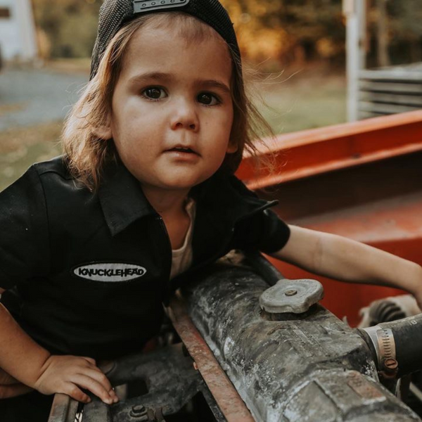 This image showcases delightful black kids' coveralls adorned with a charming Knuckleheads patch, adding a touch of whimsy to your child's wardrobe. These coveralls are not only fashionable but also comfortable, making them an ideal choice for your little one's everyday adventures.