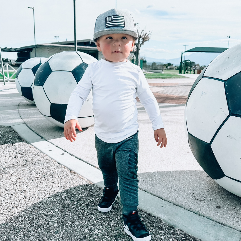 Model wearing a Grey USA Kids Trucker Hat with a black and white USA flag patch. A stylish and patriotic trucker hat designed for kids. The hat comes in a trendy grey color, featuring a striking black and white USA flag patch on the front. Elevate your child's style while proudly displaying their American spirit. Perfect for outdoor adventures and everyday wear, this hat is a fashionable and functional addition to their wardrobe.