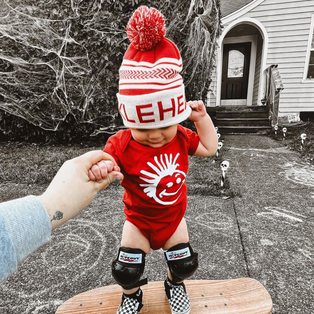 Image displaying a red and white fold-up beanie with a pom from Knuckleheads, designed for children. This versatile beanie combines a classic style with the Knuckleheads brand tag, perfect for infants and toddlers. A playful addition to the collection of Infant hats, enhancing its charm.